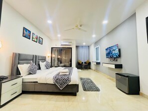 A modern bedroom with a large bed, bedside tables, a TV, and a small fridge. - Aroma Hotel Sultanpur (New Delhi)