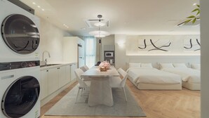 A modern kitchen with a built-in washing machine, a dining area with a table and chairs, and a bedroom with a bed and a wall-mounted TV.