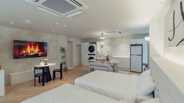 A modern hotel room with a large bed, a desk, a chair, a TV mounted on the wall, and a kitchen area in the background.