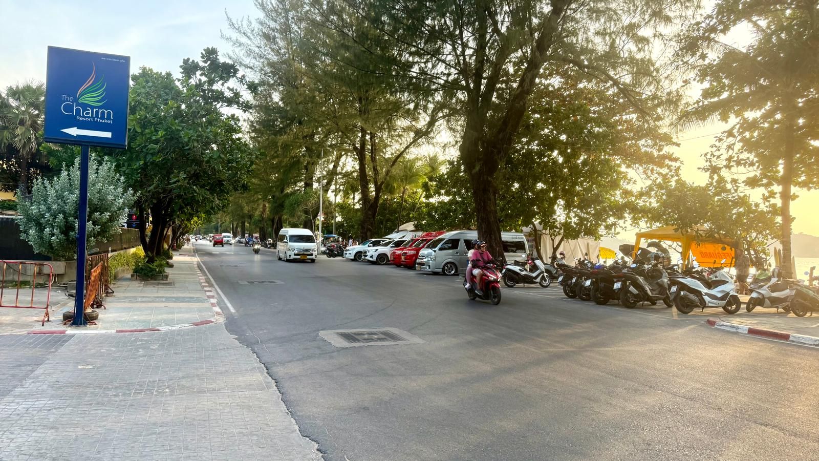 A street view with a sign for The Charm Resort Phuket, a row of parked motorcycles, and a few vehicles on the road.