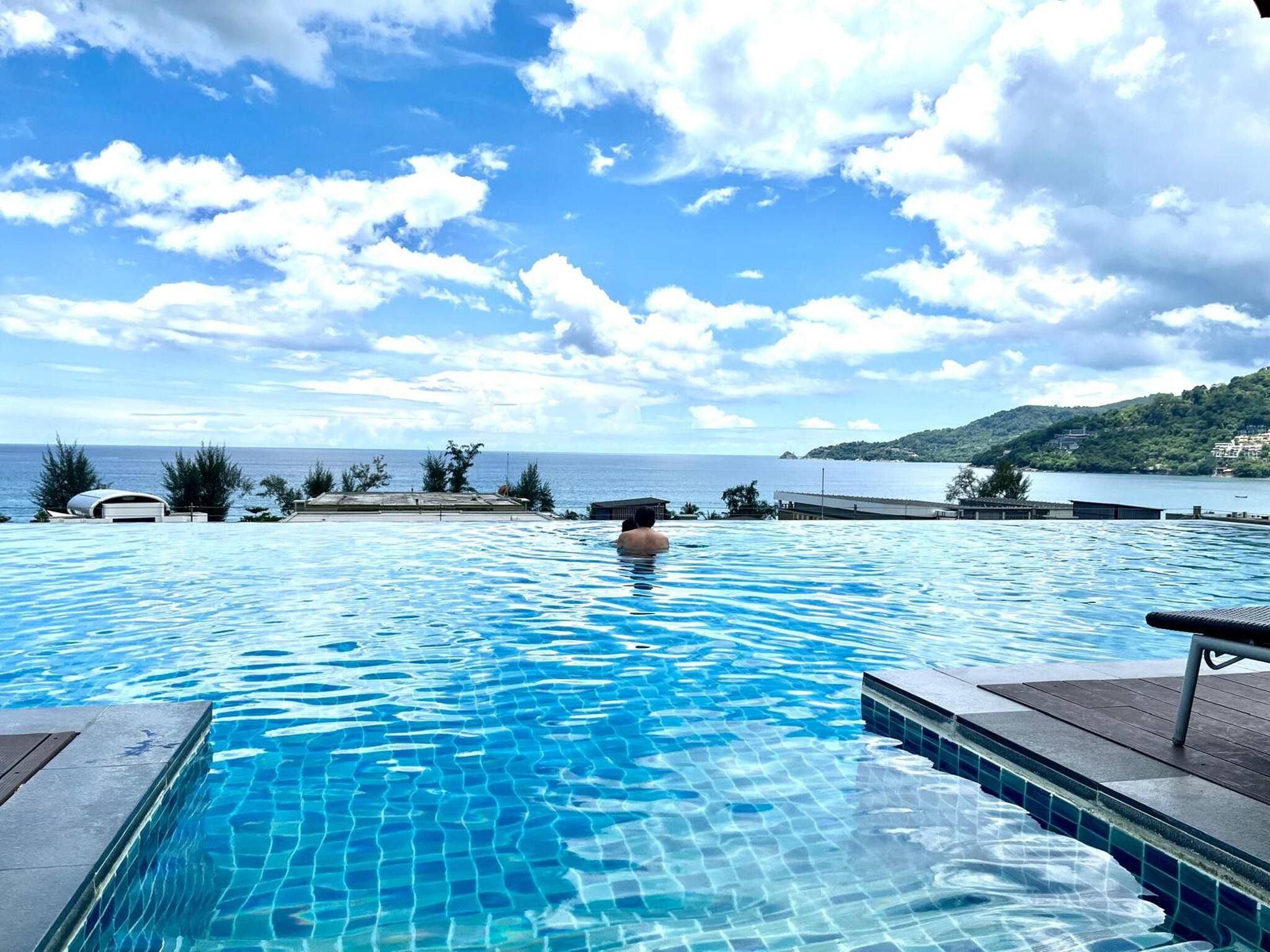 A swimming pool with clear blue water, surrounded by a deck area with a view of the sea and a hilly landscape in the background.