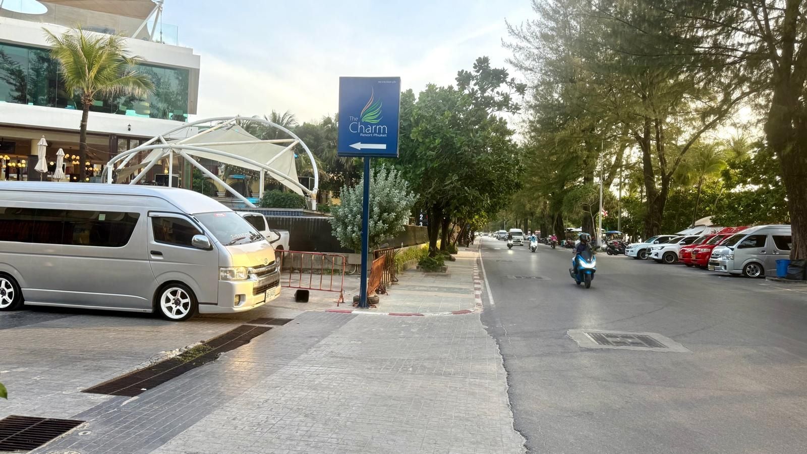 A street view with a sign for The Charm Resort Phuket, a modern building, parked cars, and a pedestrian walkway.