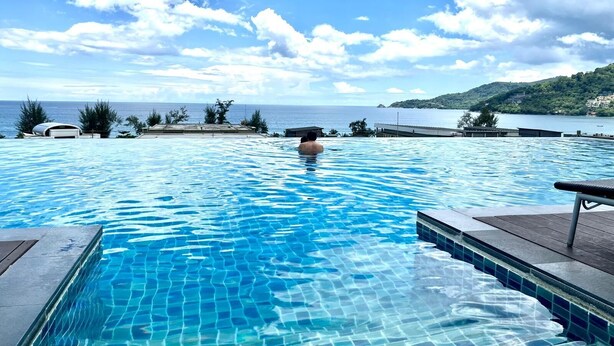 A clear blue pool with a view of the sea and a hilly landscape in the background. - Studio Apartments With Shared Pool Near Patong Beach (Patong)