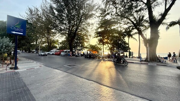A street with a sign for The Charm Resort Phuket, trees, parked vehicles, and a sunset. - Studio Apartments With Shared Pool Near Patong Beach (Patong)