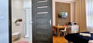 Habitación de hotel moderna con ducha, inodoro, puerta de madera, zona de comedor, televisión y cama con toallas.