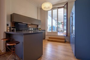 A modern kitchen with a central island, bar stools, a large window, and a staircase leading to an outdoor area.