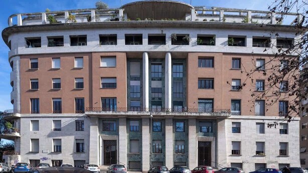 Ein mehrgeschossiges Gebäude mit einer geschwungenen Fassade, großen Fenstern und einem Balkon im oberen Bereich.