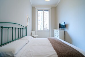 Een slaapkamer met een bed, een raam, een stoel, een dressoir en een televisietoestel.