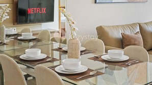 Ein moderner Essbereich mit einem Glastisch, weißen Tellern und einer Blumenvase. An der Wand ist ein Fernseher mit dem Schriftzug 'NETFLIX' angebracht.