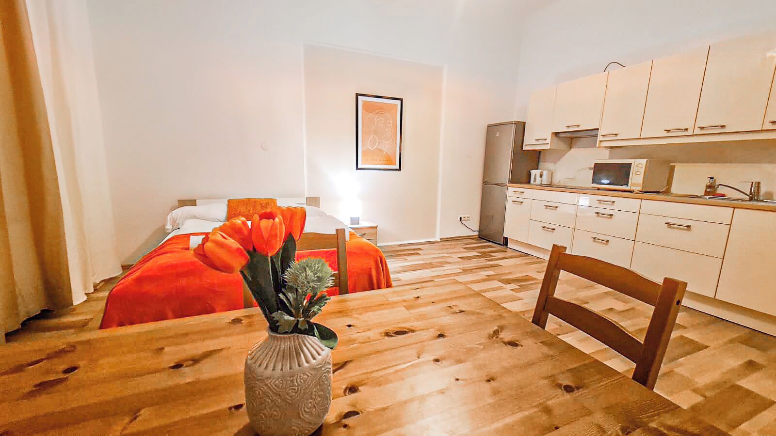 A room with a wooden floor, a bed with orange bedding, a wooden chair, a small table with a vase, and a kitchenette with white cabinets.