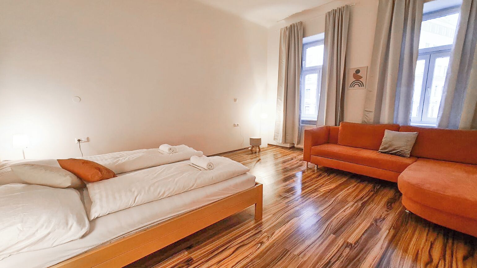 Una habitación con piso de madera, una cama con sábanas blancas, un sofá naranja y un ventanal con cortinas.