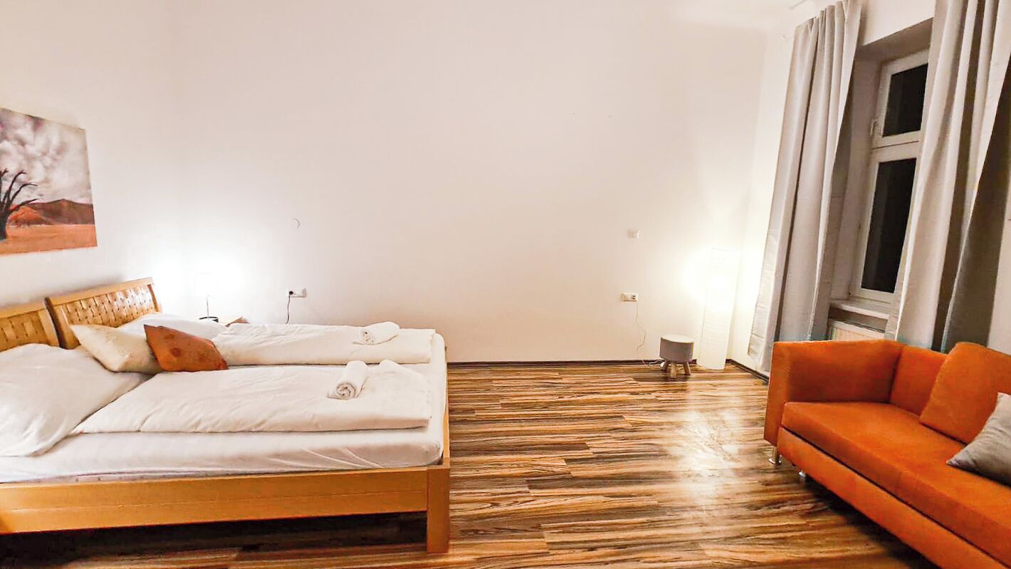A hotel room with a wooden bed, a small stool, an orange sofa, and a window with curtains.