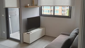 A modern living room with a sofa, a TV on a white stand, a window with blinds, and a view of buildings outside. - 2BR Apartment  T2-717 (Chalong)
