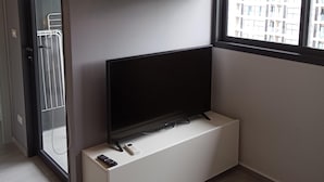 A modern living room with a flat-screen TV on a white console, a wooden shelf with decorative items, and a view of a building through a window. - 2BR Apartment  T2-717 (Chalong)