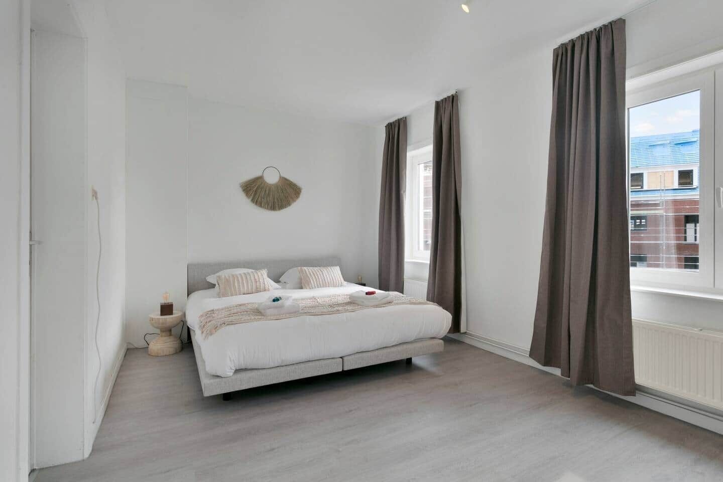 Une chambre moderne avec un grand lit, une table de chevet, un luminaire fixé au mur et une fenêtre avec des rideaux.