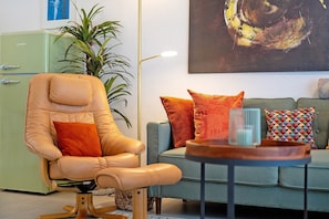 A living room with a vintage green refrigerator, a leather armchair, a blue sofa with patterned cushions, and a modern floor lamp.