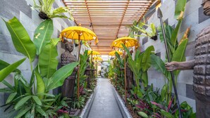 A corridor lined with large green plants and yellow umbrellas, leading to a central area with a statue. - Deluxe 2-Bedroom Tropical Haven in Seminyak with Pool & Designer Interiors (Badung)