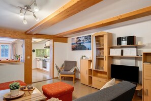 Una sala de estar con televisión, vigas de madera, un cuadro y una cocina al fondo.