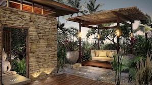 Un patio moderno al aire libre con muro de piedra, terraza de madera y zona de estar acolchada rodeada de plantas tropicales.