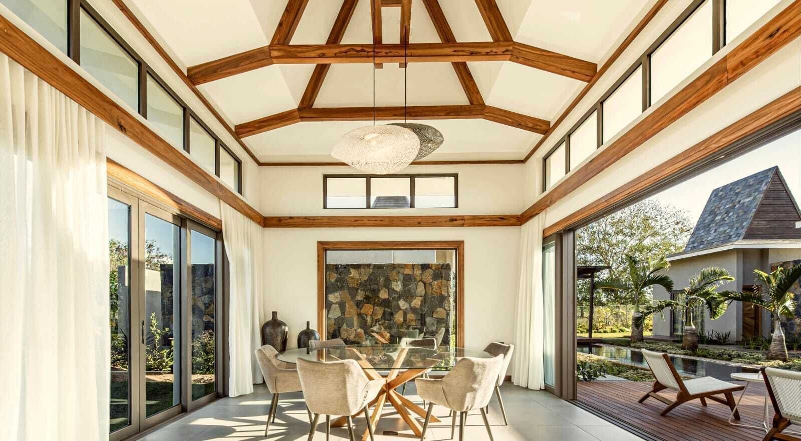 A spacious dining area with a glass table and chairs, a stone wall, and a view of a garden and pool.