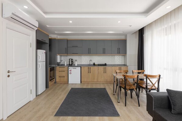 A modern kitchen with grey cabinets, a white refrigerator, and a coffee machine. There is a dining area with wooden chairs and a black sofa.