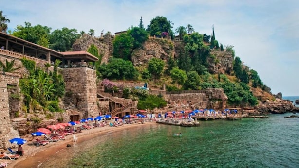 A coastal hotel with a stone facade, a beach with umbrellas, and a rocky cliff. - Moon Old Town (Antalya)