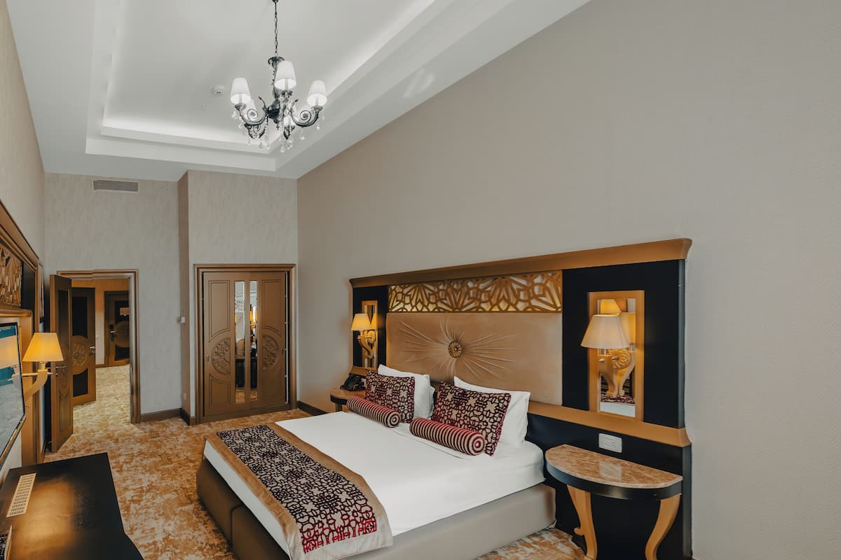 A hotel room with a large bed, a desk, a chair, a small table, and a chandelier.
