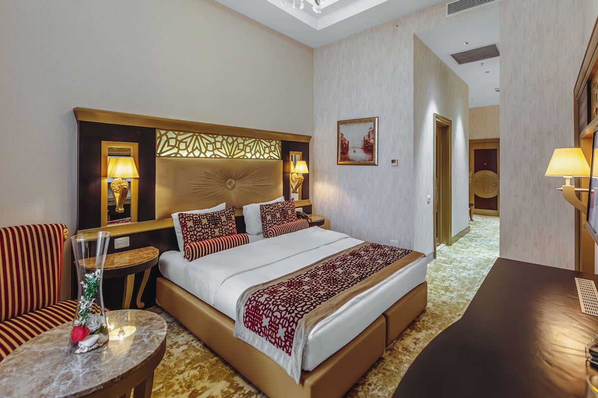 A hotel room with a large bed, a sitting area with a chair, a small table with a vase, and a nightstand with a lamp.