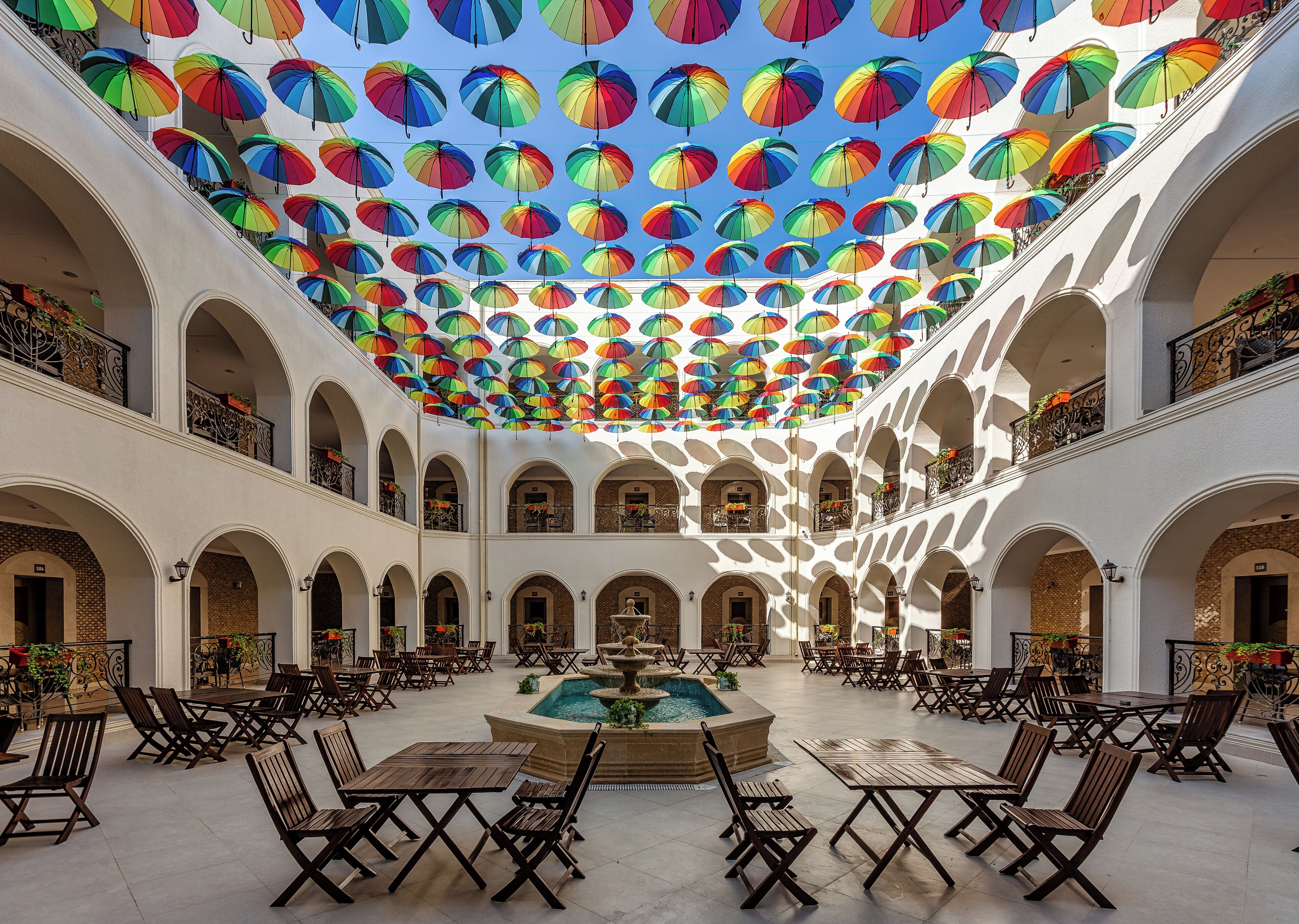 A spacious courtyard with a central fountain, surrounded by wooden tables and chairs, and a ceiling adorned with numerous colorful umbrellas.