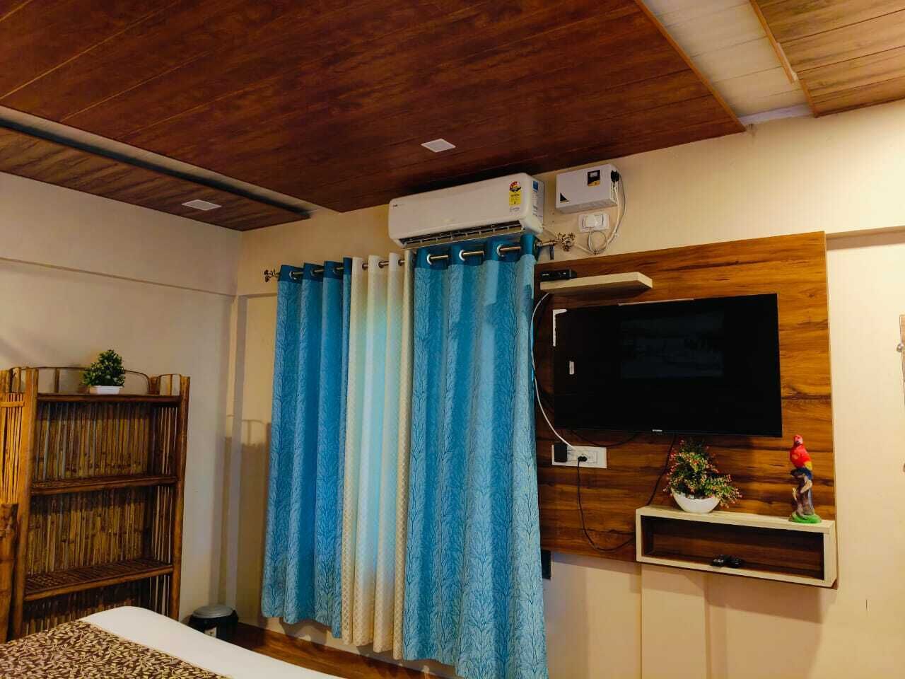 A bedroom with a wooden ceiling, a mounted TV, a bed with a patterned bedspread, and a window with blue curtains.