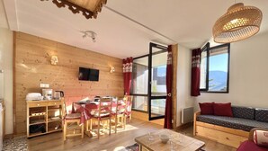 Une pièce chaleureuse avec une table à manger en bois, des chaises, un canapé et une table basse. Un téléviseur est fixé au mur et il y a une fenêtre avec des rideaux.