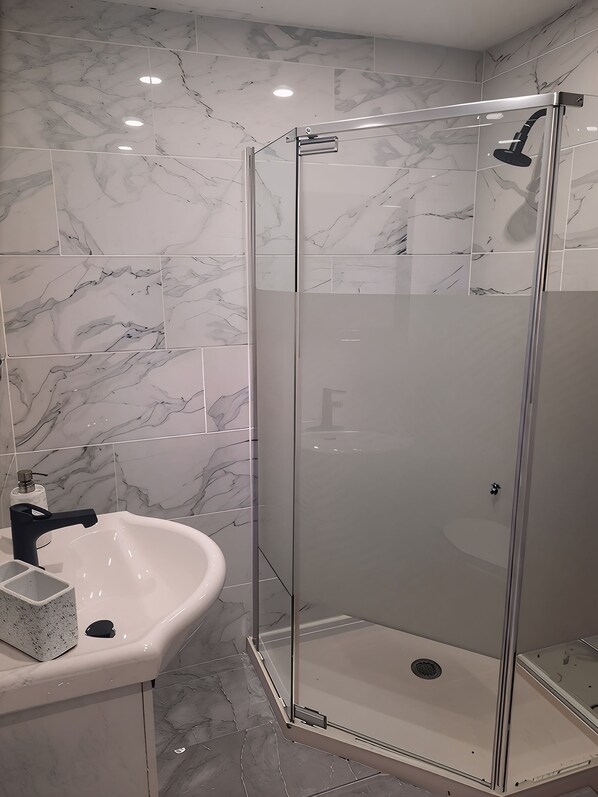 Un baño con ducha envuelta en vidrio, un lavamanos blanco y baldosas con diseño de mármol.