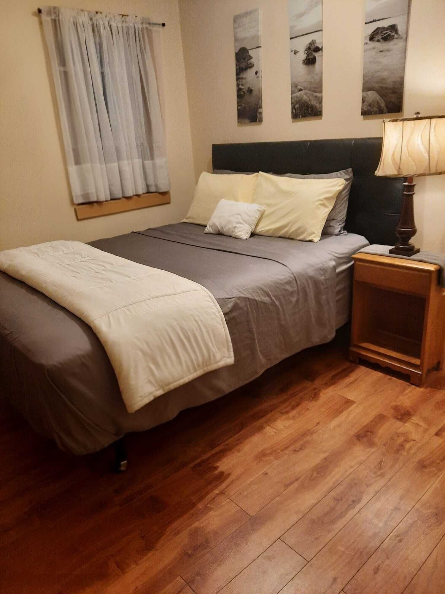 Un lit bien fait, avec une couette grise et blanche, une table de chevet en bois sur laquelle se trouve une lampe, et une fenêtre avec des rideaux.