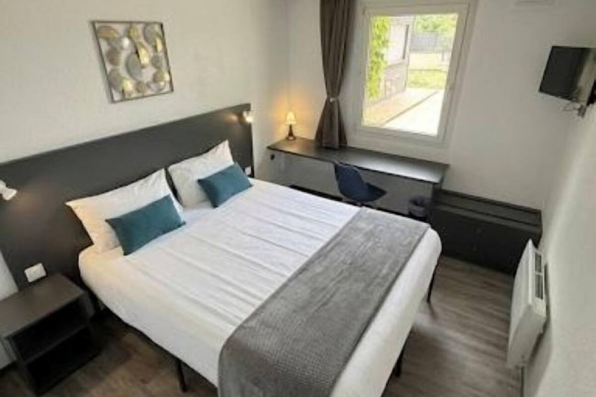 Hotellihuone, jossa on sänky, yöpöydät, työpöytä, tuoli, ikkuna verhoilla, seinälle asennettu televisio ja patteri.
