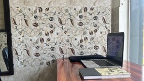 A wooden desk with a laptop and a book, a black chair, and a decorative wall with a leaf pattern. - The Harikanta Residency (New Delhi)