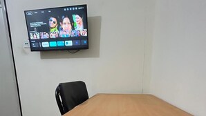 A conference room with a wooden table and black chairs, a flat-screen TV mounted on the wall displaying a social media app, and a tiled floor. - The Harikanta Residency (New Delhi)