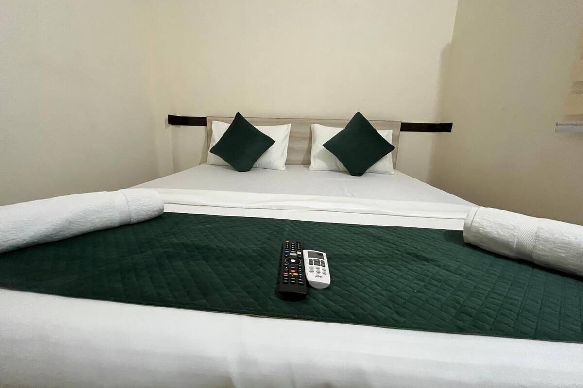 Una cama bien hecha con una colcha verde, dos almohadas y dos controles remotos sobre la cama.