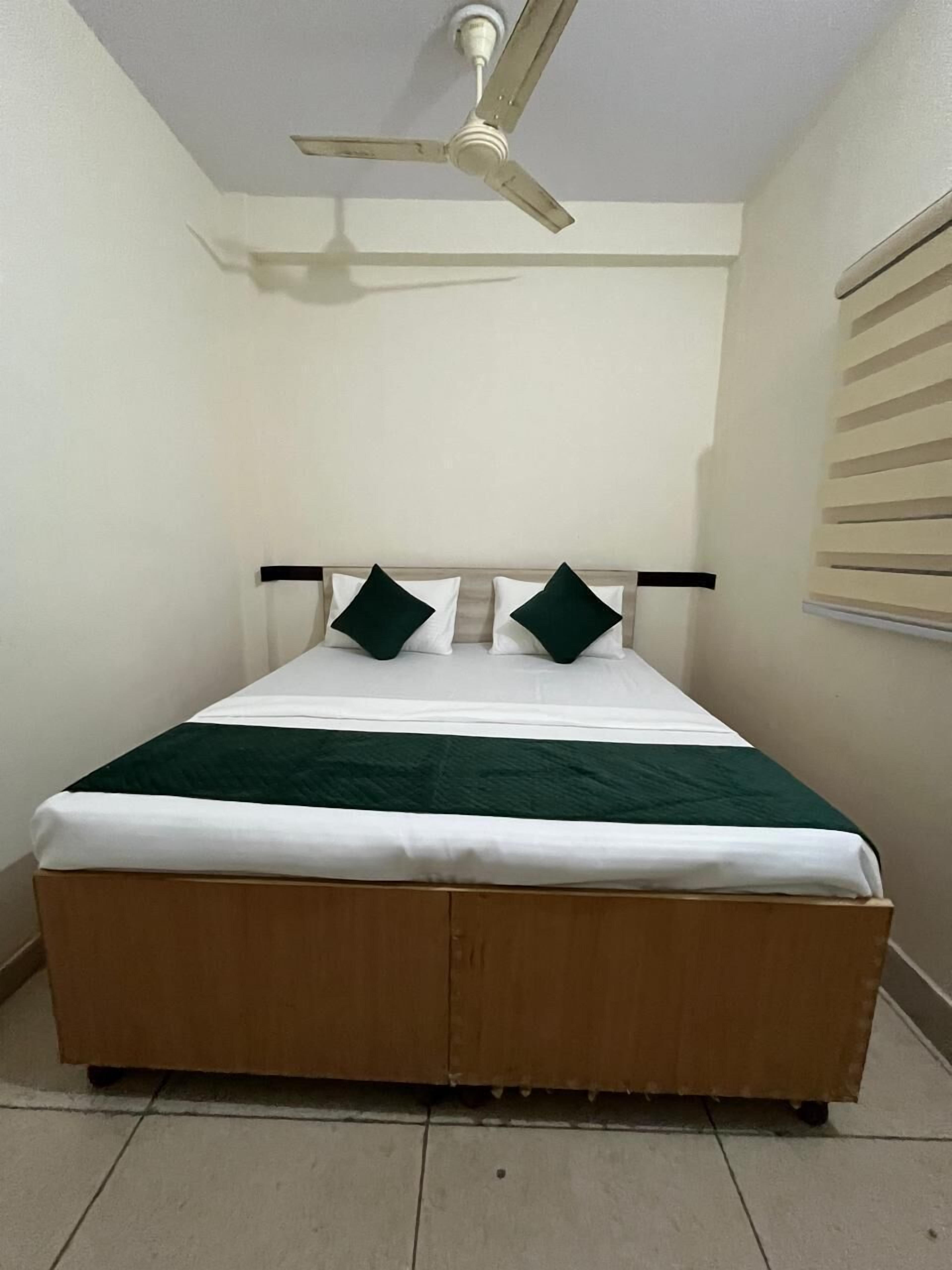 Een enkel bed met houten frame, wit beddengoed en twee groene kussens. Boven het bed is een plafondventilator bevestigd.