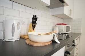 A kitchen with white cabinets, a white kettle, a microwave, and a wooden cutting board with a bowl and a wooden spoon. - 1C Lime Street Apartment (Bedford)