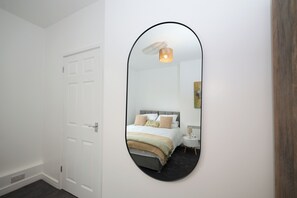 A bedroom with a white door, a round mirror, a bed with pillows, a bedside table, and a lamp. - 1C Lime Street Apartment (Bedford)