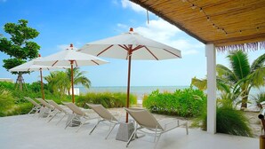 A beachside lounge area with white lounge chairs and umbrellas, a covered seating area, and a view of the ocean.