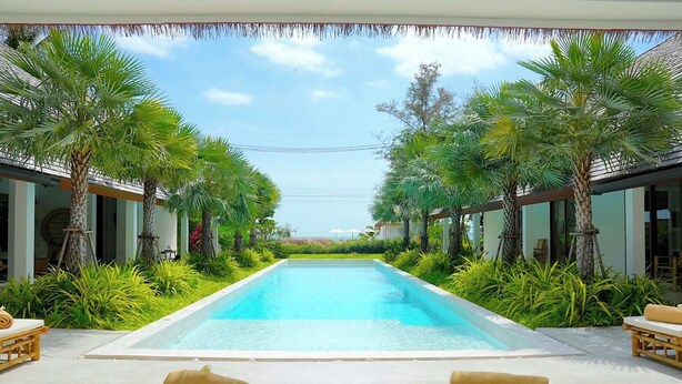 A rectangular swimming pool with a clear blue water surface, surrounded by lush greenery and palm trees. There are lounge chairs with yellow towels placed on the poolside.
