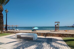 Ein Weg direkt am Strand mit einer Bank, Sonnenschirmen und einem Rettungsschwimmerstand.