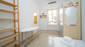 A hotel room with a bed, a small table, a wooden shelf, a wardrobe, and a door leading to another room.