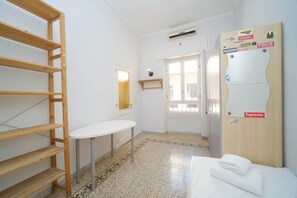 A hotel room with a bed, a small table, a wooden shelf, a wardrobe, and a door leading to another room.