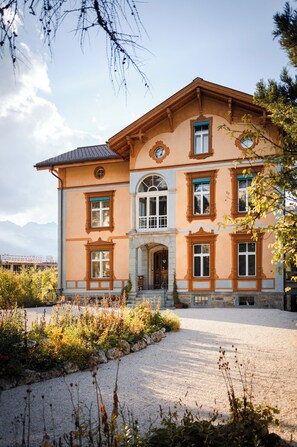 A large, two-story building with a symmetrical facade, featuring a central entrance with a stone archway, multiple windows with decorative frames, and a gravel driveway leading to the entrance. - Villa Klainguti (Pontresina)