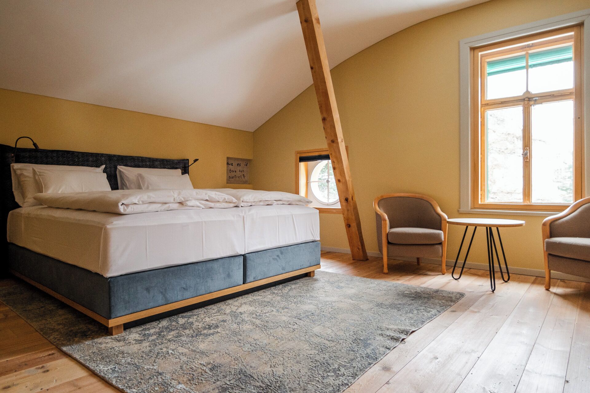 Ein Schlafzimmer mit einem Bett, zwei Stühlen, einem kleinen Tisch, einem Fenster und einem Holzbalken.