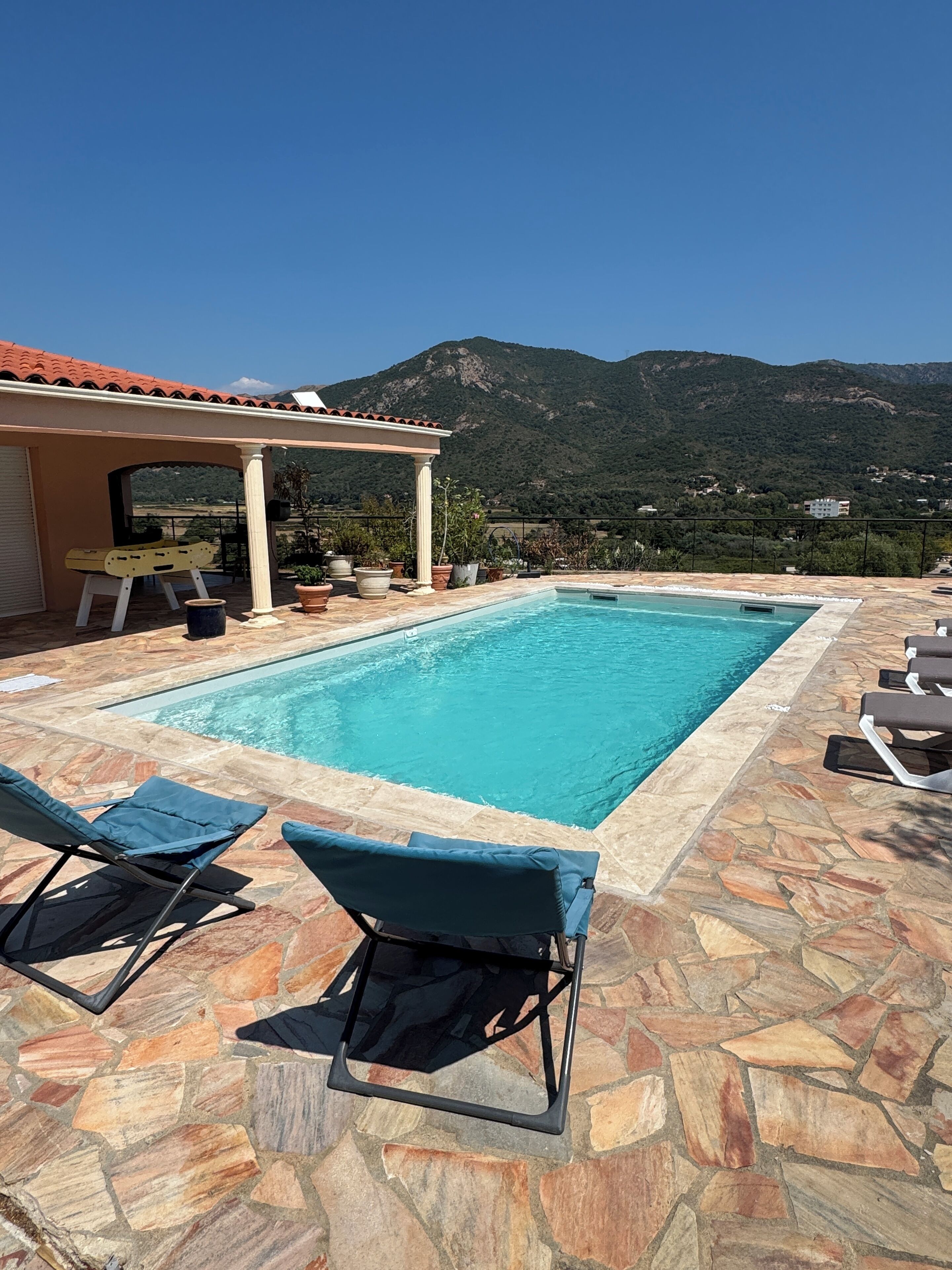 A swimming pool with stone tiles, two lounge chairs, a pergola, and a mountain view.