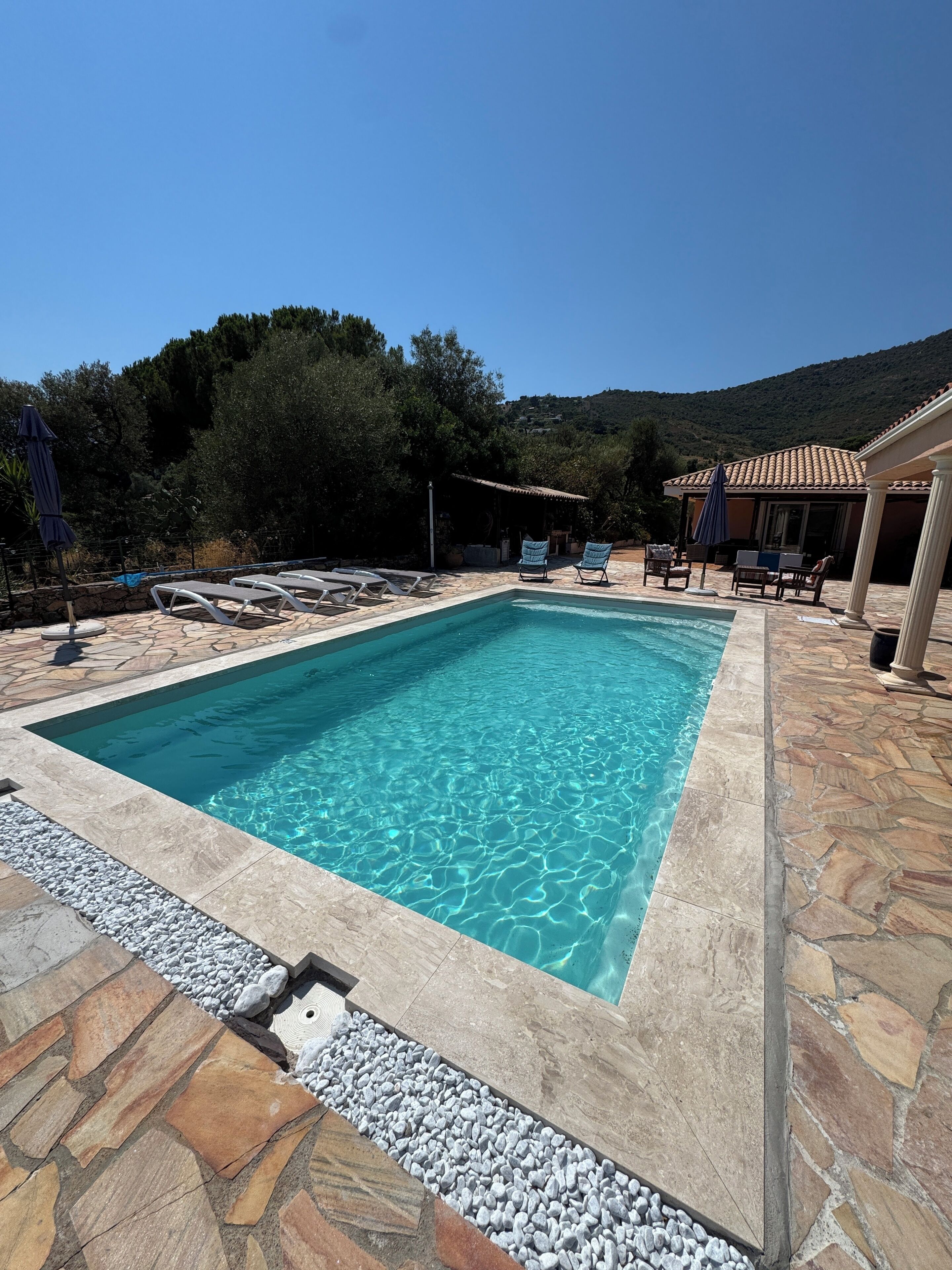 A rectangular swimming pool with clear blue water, surrounded by stone tiles and a pebble border. There are lounge chairs and umbrellas around the pool area.