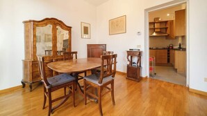 Een eetruimte met een houten tafel en stoelen, een houten kast, en een keuken zichtbaar door een open doorgang.
